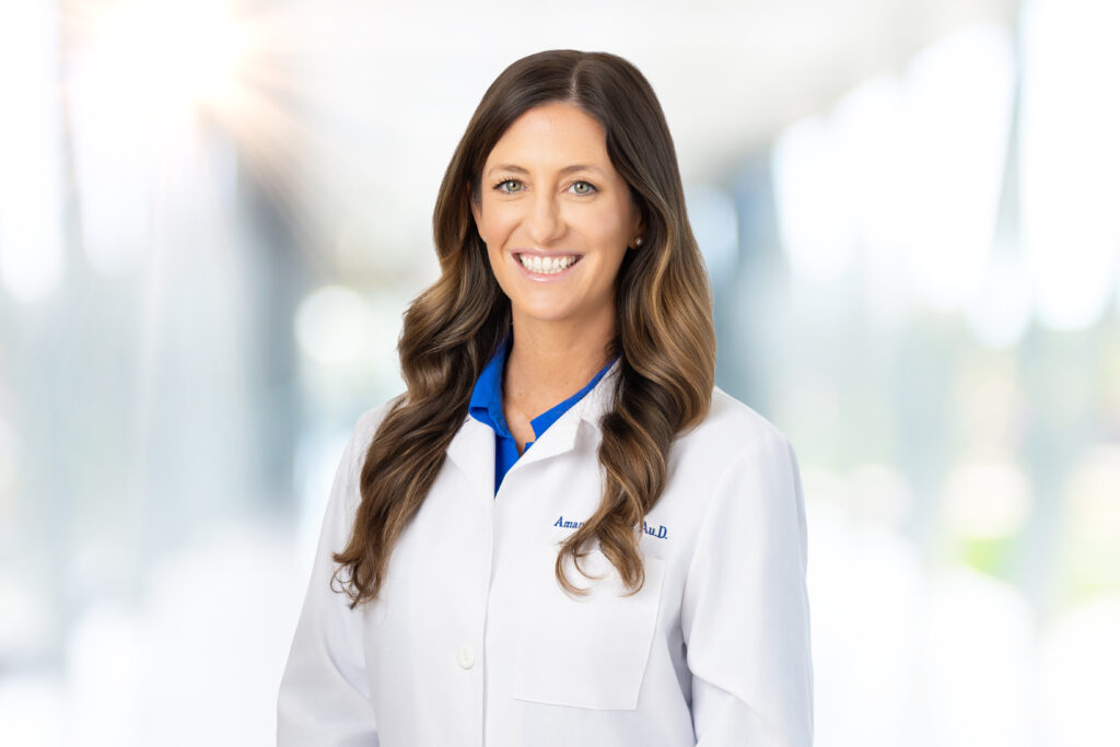 A woman with long brown hair wearing a white lab coat and blue shirt smiles at the camera. The background is bright and blurred, suggesting a professional or medical setting.