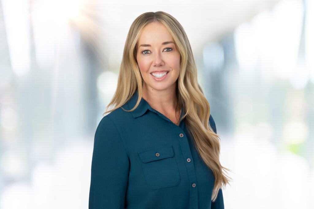 A woman with long blonde hair, wearing a teal button-up shirt, smiles at the camera. The background is blurred and brightly lit, creating a soft and professional atmosphere.