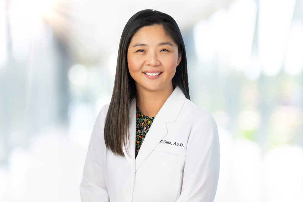 A woman with straight dark hair, wearing a white lab coat and a floral top, smiles at the camera in a bright, softly blurred indoor setting.