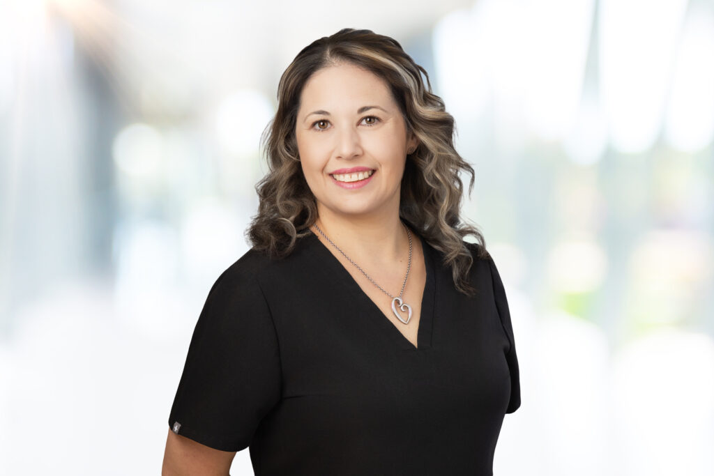A woman with wavy brown hair wearing a black top and a heart-shaped necklace smiles at the camera, standing in front of a blurred, bright background.