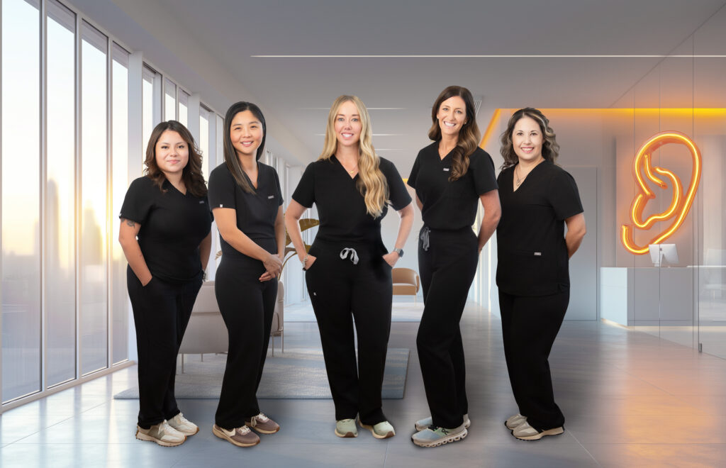Five women in black scrubs stand smiling in a modern, bright medical office with large windows and a neon ear sign, ready to help patients address hearing loss with expert care.