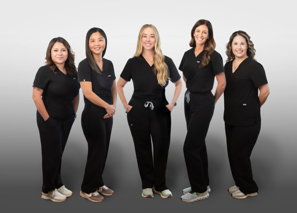 Five women wearing black medical scrubs and sneakers stand side by side, smiling at the camera against a light gray background.