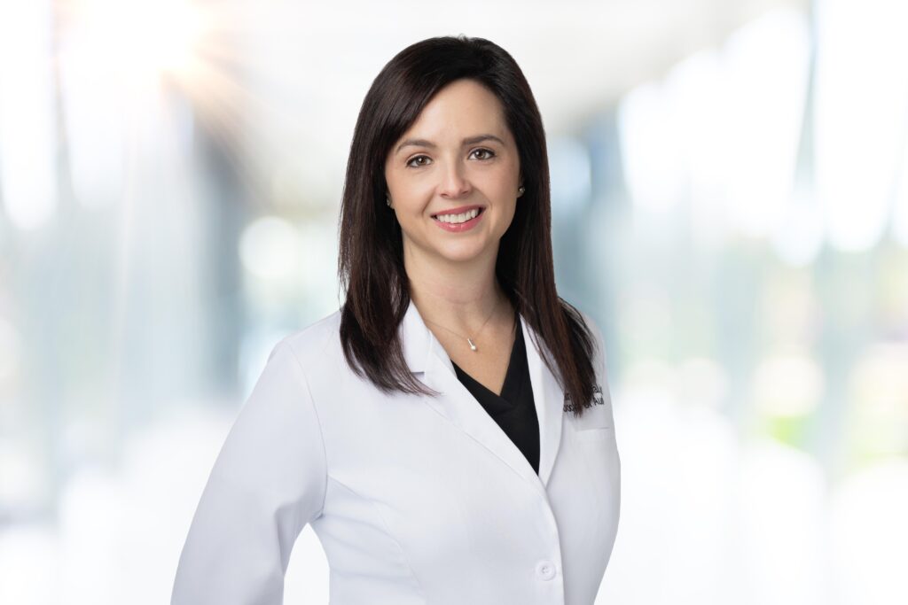 A woman with straight dark hair, wearing a white lab coat over a black top, stands smiling in a bright, blurred indoor setting. The background has soft light and indistinct shapes.