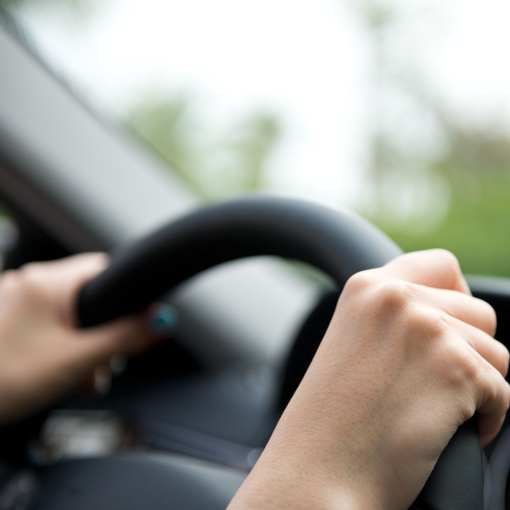 Close-up of a person’s hands gripping a steering wheel inside a car, with a blurred view outside the window suggesting movement or focus on driving.