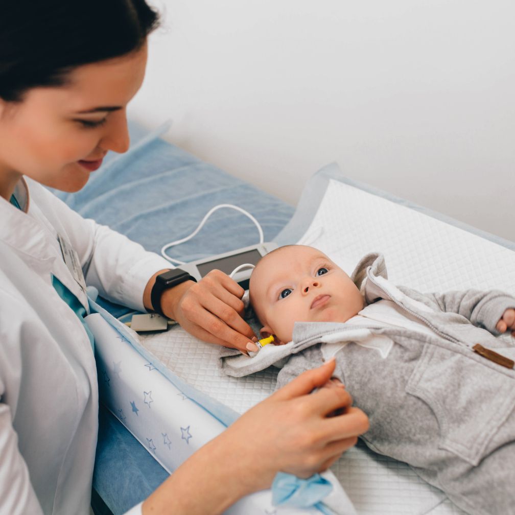 A healthcare professional checks a babys ear with a device as the baby lies on an exam table, looking up. The scene appears to be in a medical office or clinic.