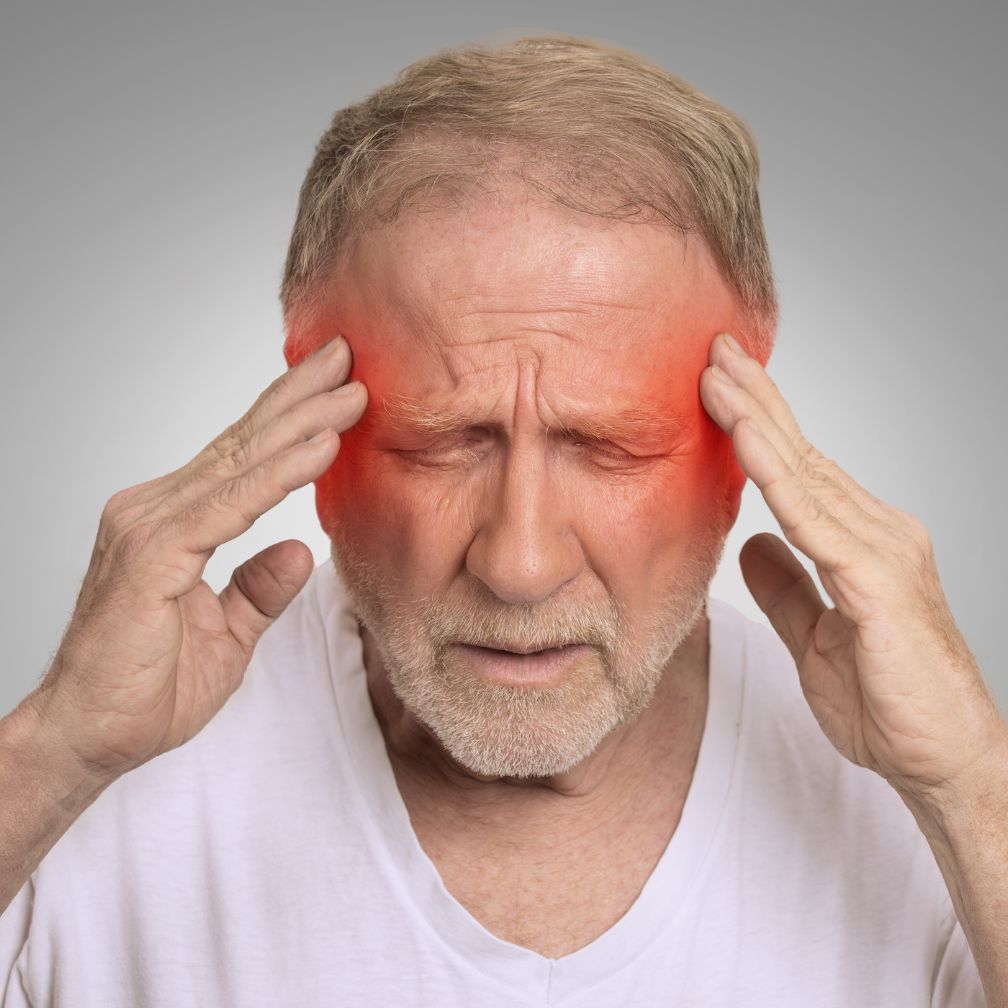 An older man holds his head with both hands, eyes closed in pain. Red shading highlights his temples, suggesting a headache or migraine.