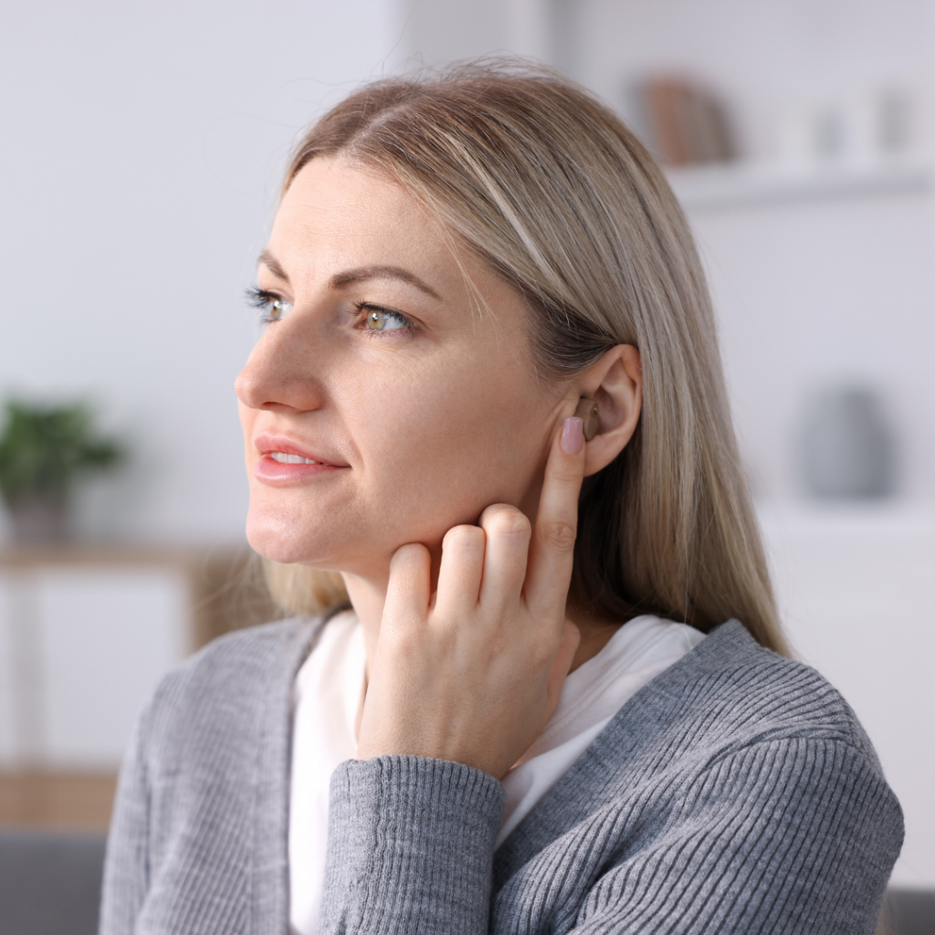 A woman with blonde hair touches her ear while looking to the side, wearing a gray sweater and sitting indoors with a soft, blurred background.