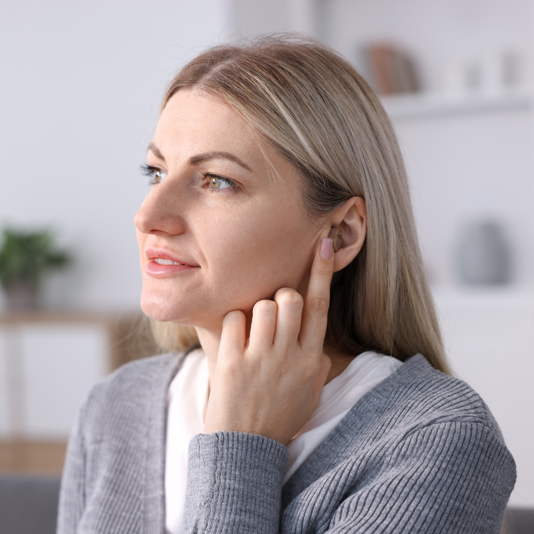 A woman with blonde hair touches her ear while looking to the side, wearing a gray sweater and sitting indoors with a soft, blurred background.